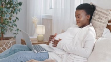 Afro-Amerikan anne adayı zenci kadın hamile karnını okşuyor hamileliğin tadını çıkarıyor modern dizüstü bilgisayar uygulaması internet servis tarayıcıları ile internetten mal sipariş ediyor.