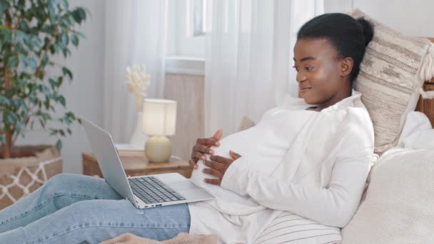 妊娠を楽しんで妊娠中の腹を撫でるベッドの上に横たわるアフリカ系アメリカ人の期待の母親黒人民族の女性は、現代のラップトップコンピュータアプリのWebサービスを使用しています遠隔操作で商品をオンラインで注文 — ストック動画