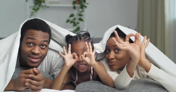 Afrikansk amerikansk etnisk sort blandet race familie har det sjovt indendørs liggende på sofaen dækket med tæppe gør sjove ansigter viser kikkert form med fingrene som briller gestus smilende ser på kameraet – Stock-video