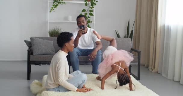 Afro-Amerikaanse familie tijd doorbrengen thuis, Afrikaanse vader zit op de bank op zoek naar mobiele telefoon, zwarte etnische moeder speelt met meisje dochter in roze jurk acrobatische poses doen op de vloer op zacht tapijt — Stockvideo
