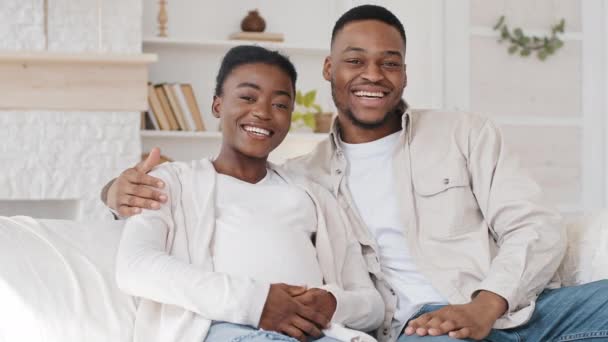 Africano casal família pais afro preto étnico marido e mulher acenando mão Olá saudação falando sobre gravidez olhando para câmera sorrindo acariciando barriga grávida fazendo videochamada da sala de estar — Vídeo de Stock