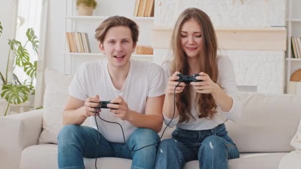 Caucasian married couple Millennial boyfriend and girlfriend woman and man, friends sitting on couch playing video game using console controllers enjoying competition girl wins making victory gesture — Stock Video