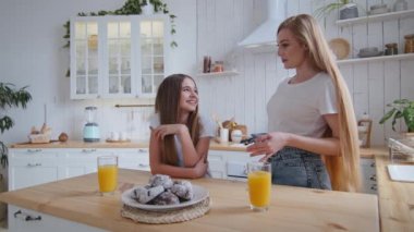 Kafkasyalı yetişkin anne ve küçük kız çocuk okul kızı ellerinde kurabiyelerle ve portakal suyu bardaklarıyla yemek masasının yanında durmuş şakalaşarak eğleniyorlar.