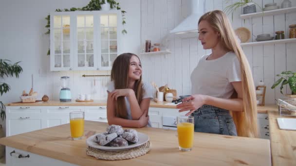 Kafkasyalı yetişkin anne ve küçük kız çocuk okul kızı ellerinde kurabiyelerle ve portakal suyu bardaklarıyla yemek masasının yanında durmuş şakalaşarak eğleniyorlar. — Stok video