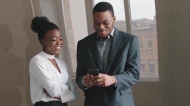 Mutlu Afrikalı Amerikalı stajyerler telefona, akıllı telefon cihazına bakıp gülüyor, mesajları kontrol ediyor, medyayı okuyor, iş saatlerinde dinleniyor. E-ticaret kullanıcısı, yeni uygulama, modern teknoloji ile uzaktan iletişim.
