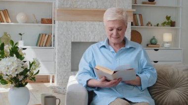 Gerçekten mutlu, gülümseyen yetişkin bir bayan oturma odasında kanepede oturuyor. Orta yaşlı kadın elinde kitap tutarak evde yalnız başına modern roman edebiyatıyla vakit geçirmekten zevk alıyor.