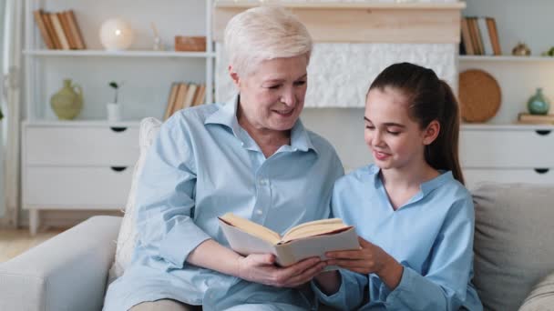 Starsza babcia trzyma książkę i dziewczynka siedząca razem na kanapie w domu. Szczęśliwa babcia uczy wnuka czytania w domu. Uśmiechnięta starsza pani, dumna z mądrej wnuczki. — Wideo stockowe