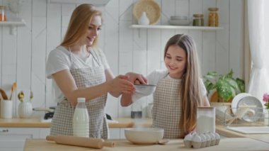 Yetişkin anne, sarışın kadın ve küçük kız önlük takıyor. Korkak buğday unu pişirmeyi öğreniyorlar. Demir elek kullanıyorlar.