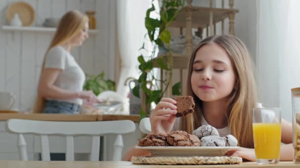 Klein meisje tiener kind dochter schoolmeisje zitten aan tafel in huis keuken eten heerlijke zelfgemaakte chocolade koekjes genieten van het ontbijt op de achtergrond van wazig moeder afwassen bord — Stockvideo