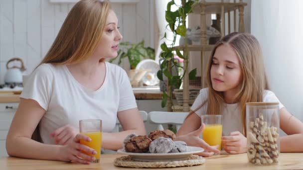 Mor och dotter tonåring barn flicka sitter tillsammans vid bordet i köket dricker naturlig apelsin tropisk frisk juice färsk i glasögon talar vid frukost på morgonen. Mamma strokes baby på huvudet — Stockvideo