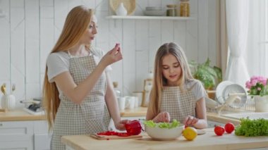 Yetişkin anne ev hanımı ve kız çocuğu yardımcısı mutfak önlüğü giyer taze doğal salata için sebze malzemesi keser anne kırmızı biber yer çocuğu beslemeyi dener