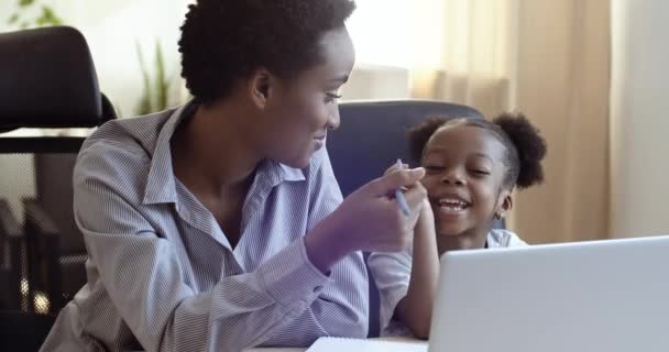 Afrikansk sort kvinde mor hjælper sød elskede datter skolepige med lektier sidder sammen på laptop studie online i netværk skrive på papir tage noter i notesbog, hjem undervisning læringskoncept – Stock-video