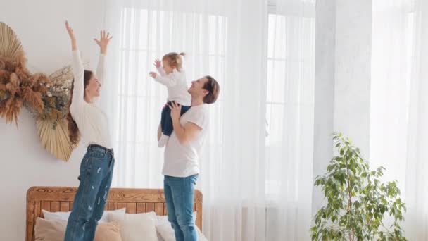 Pais jovem casal caucasiano família pai segurando pequena filha bebê menina levanta-se no ar mãe levanta as mãos criança criança repete, mãe e pai sorriso desfrutar feliz fim de semana em casa quarto na cama — Vídeo de Stock