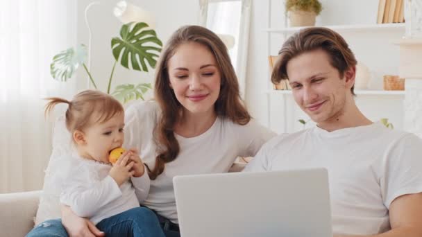 Портрет кавказької родини використовує ноутбук, що сидить на дивані вдома. Молода мати тримає дитину-дочку дівчинку, яка їсть яблуко, розмовляє з батьком-чоловіком, дивлячись на покупки на екрані комп'ютера онлайн — стокове відео