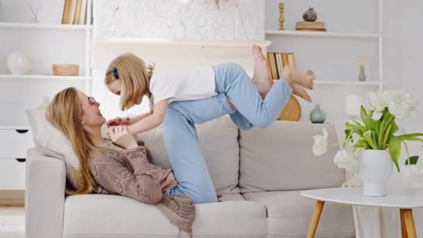 Мать-блондинка держит поднимает маленькую дочь девочка ребенок на ногах в воздухе, делая акробатические упражнения баланс лежа на диване дома в гостиной весело играть в игры притворяются самолета крытый — стоковое видео