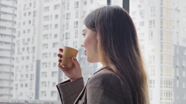 Caucasian young millennial business woman leader boss specialist professional stands in office looking out window at skyscraper city drinks tea coffee from cardboard cup turns head looks into camera — Stock Video