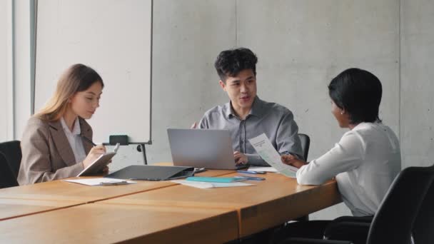 Liderul sefului de afaceri asiatic care sta la masa cu laptopul discuta despre startup-ul strategiei de proiect cu colegii de sex feminin femeie caucaziana si femeie de afaceri africana care scrie in notebook, echipa multietnica — Videoclip de stoc