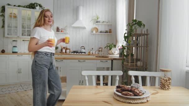 Caucásico hermosa mujer madre ama de casa con el pelo largo lleva vaqueros preparándose para la cena de desayuno pone en la mesa dos vasos con jugo de naranja tropical fresco y galletas de chocolate en la cocina — Vídeos de Stock