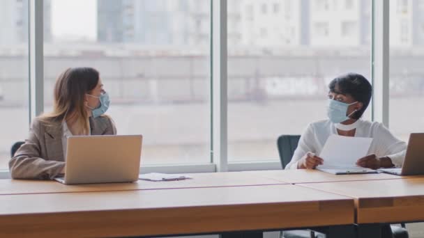 Zwei multiethnische Arbeiterinnen, eine kaukasische Managerin und eine afrikanische Geschäftsfrau, die im Büro am Tisch sitzen, halten Abstand und tragen medizinische Schutzmasken von der Arbeit mit Laptops. — Stockvideo