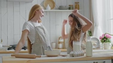 Yetişkin güzel anne, sarışın ev hanımı anne ve kız çocuğu önlük giyiyorlar mutfakta birlikte yemek pişiriyorlar, liseli kız başarı atlayışı yapıyor eğleniyor zafer jestleri yapıyor.