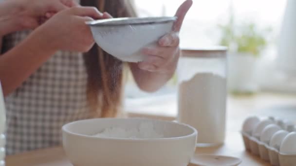 Закрыть женские и детские руки держит железо просеивает пшеничную муку в миску приготовления теста для выпечки белого хлеба печенья. Неузнаваемая дочь помогает матери с приготовлением ингредиентов — стоковое видео