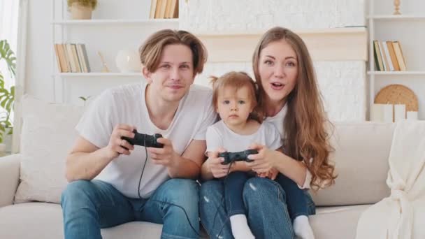 Кавказская семья играет в видеоигры консоли дома сидя на диване молодая мать и маленькая дочь ребенок маленькая девочка выиграть конкурс проигравший отец смешной досуг, пара с ребенком наслаждаться видеоиграми — стоковое видео