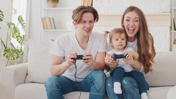 캅카스 청년층의 젊은 가족 아버지와 아름다운 어머니가 소파에 앉아 비디오 게임 콘솔을 하고, 엄마는 어린 딸을 안고 비디오 게이밍 박수를 받으며 환호하고 있습니다. — 비디오
