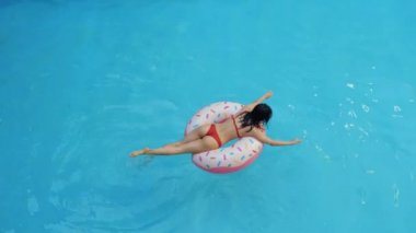 Sıska genç kız kırmızı bikini giyiyor, şişme halka üzerine uzanmış mavi suda kürek çekiyor, dalgayı elleriyle kesiyor, bacakları suyun altında hareket ettiriyor, su sıçratıyor, havuzda yüzüyor, tepeden bakıyor.