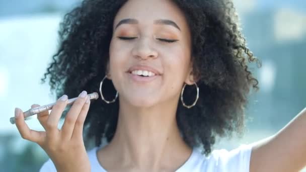 Hermosa chica africana con maquillaje, peinado afro está grabando mensaje de voz en el teléfono, hablando con su amigo, riendo adorable de broma. Estudiante americano étnico se comunica, tiene buen tiempo al aire libre — Vídeo de stock