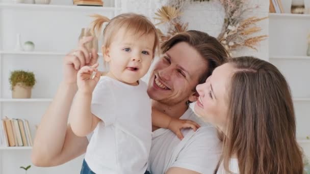 Portrét kavkazské rodiny mladý rodič drží malé dítě dcera batole mluví objímání s úsměvem mazlící se dítě dívka ukazující směr s prstem na stranu v okně maminka a tatínek odvrací pohled — Stock video
