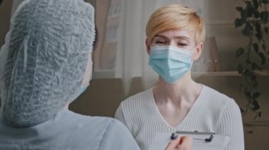 View from behind unrecognizable woman doctor specialist practitioner medical worker consults mature sick female patient in protective mask asks about symptoms disease writes down diagnosis in hospital