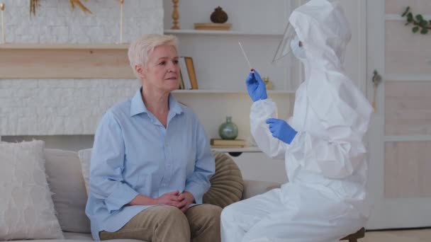 Female doctor nurse medical worker in special protective uniform makes test PCR takes sample saliva analysis from old woman grandmother of elderly lady at home during covid-19 coronavirus epidemic — Wideo stockowe