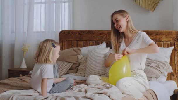 Caucasian family lonely mother and little girl child sitting on bed in bedroom talking laughing playing mom ties yellow balloon gives to baby communicating, concept of holiday celebration at home