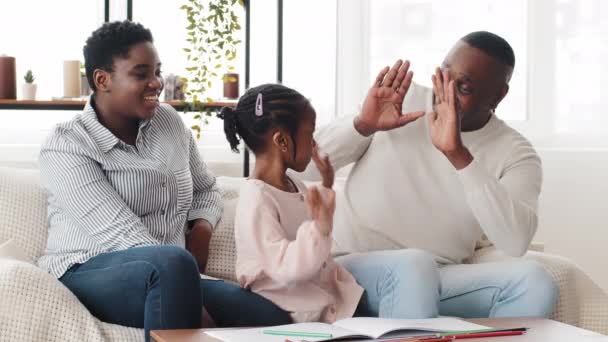 Glad afrikansk familie afro amerikanske forældre hjælper sort lille pige skolepige barn tegne billede lektier sidder på sofaen ved bordet baby giver high five til mor og far vellykket teamwork gestus – Stock-video