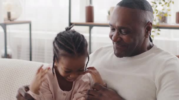 Glad afrikansk amerikansk familj mogen man med rynkor far farfar med liten dotter flicka barn sitter tillsammans hemma på soffan talar skrattar kommunicerande samtal hålla hand — Stockvideo