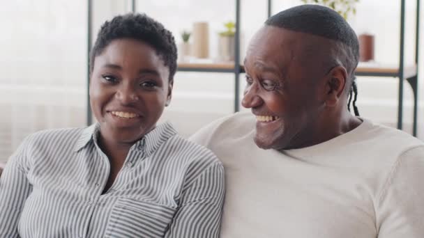 Retrato de família casal africano diferentes idades afro jovem e maduro homem étnico negro sentado no sofá falando rindo. Pai sênior e filha de raça mista olhando para câmera conversando abraços — Vídeo de Stock