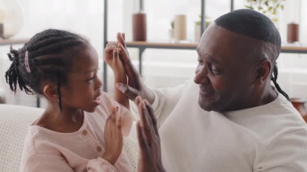 Afro américaine petite fille presse sur les mains paumes touche mature père grand-père africain noir homme bébé cris fait effort querelle papa joue avec enfant rire sourire avoir du plaisir sur le canapé — Video