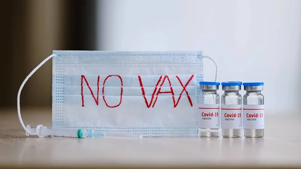 Close-up transparent glass bottles with liquid vaccine medicine anti-covid-19 coronavirus on table near syringe and medical mask with inscription no vaccination, refusing immunization during pandemic