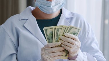 Yakın plan tanınmayan doktor hemşire, doktor, cerrah, beyaz önlüklü, koruyucu yüz maskeli, lateks eldivenli, yığınla para tutan banknotlar, klinikte rüşvet alan banknotlar.