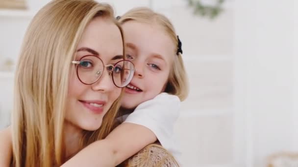 Portrett av en lykkelig ung mor i briller med datter som klemmer hjemme og smiler mens hun knytter bånd. Lille jentebarnehage babyunge omfavne klem elsket mor ved nakke kose – stockvideo