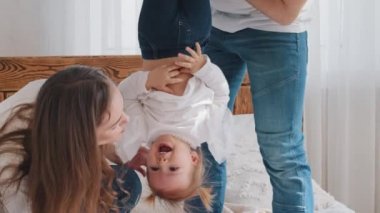 Yakın plan, güçlü bir baba kucağında küçük bir bebek bebek ters asılı kız çocuğu akrobatik poz veriyor mutlu anne kahkahalar atıyor, beyaz aile yatakta oynuyor.