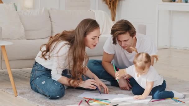 Os pais jovens ajudam a filha pequena criança pré-escolar menina desenhar quadro com lápis de cor sentado no chão. Bebê criança desenho mãe aplaude apoio, família criativo lazer ativo em casa — Vídeo de Stock