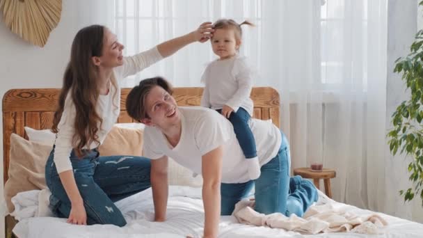 Kaukasisk lycklig familj har roligt tillbringar helgen tillsammans på sängen, glad pappa håller liten dotter på rygg piggyback rida spel leka med liten flicka barn skrattar, omtänksam mamma ler — Stockvideo