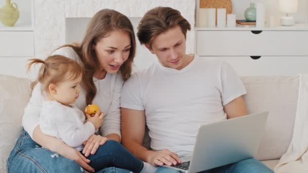 Famiglia caucasica giovane madre padre millenario acquirenti affittuari utenti con figlia piccola bambina seduta sul divano a casa utilizzando laptop guarda lo schermo cerca nuovo appartamento shopping online navigazione — Video Stock