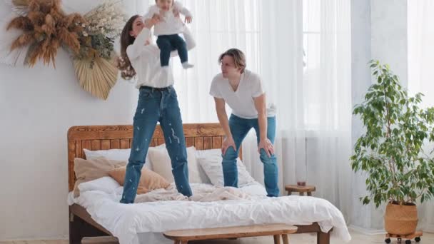 Caucasico giovane famiglia genitori divertirsi a casa in camera da letto in piedi sul letto, forte adulto madre mamma sollevamento bambino figlia bambina in aria gettando alto bambino volare padre applaude sostiene — Video Stock