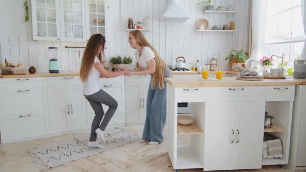 Caucasian adult active mother babysitter with long hair and teenager girl daughter child in jeans holding hands dancing making same legs movements enjoy dance in kitchen at home having fun together — Stock Video