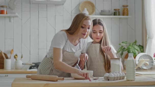 Dorosła matka z długimi włosami i córka dziewczynka gotowanie ciasta razem w domu w kuchni stojąc przy stole, mama rysuje kształt serca z palcem na mące, uczennica dotyka mamusi przez rozmazy z nosa — Wideo stockowe