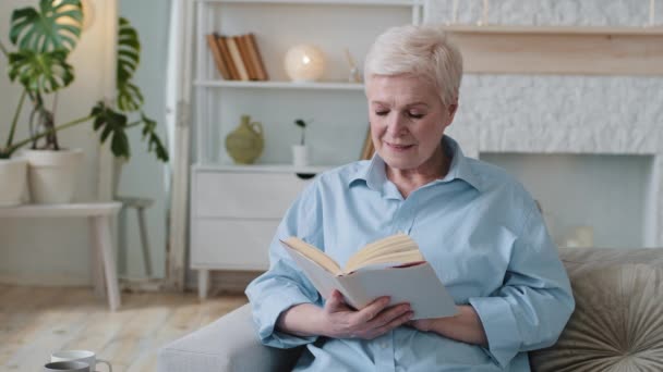Entspannt liest eine alte Frau einen Bestseller aus dem Buch, sitzt auf dem Sofa und verbringt das gemütliche Wochenende zu Hause. Glückliche Seniorin genießt Freizeit mit Hobby und ruht sich auf Couch im Wohnzimmer aus. — Stockvideo