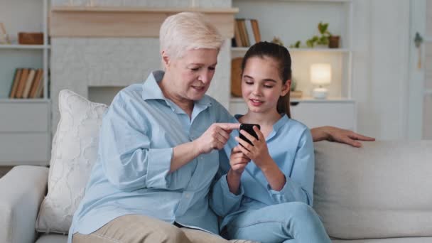 Boldog érett nagymama és kis aranyos unoka okostelefon alkalmazásokat használ a kanapén. Szülői felügyelet biztonsága gyerek az interneten, szórakoztató és oktatási mobil app felhasználók. Otthoni szabadidő modern technológiai koncepcióval — Stock videók