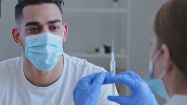 Невизначена жінка-лікар в рукавичках тримає шприц з вакцинами, готуючись зробити щеплення від кованого коронавірусу-19 до хворого арабського пацієнта в медичній масці крупним планом — стокове відео